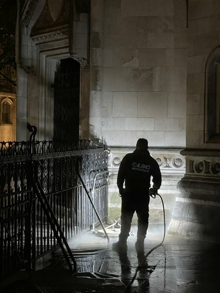 A man with a hose conducting exterior cleaning on a commercial building at night.