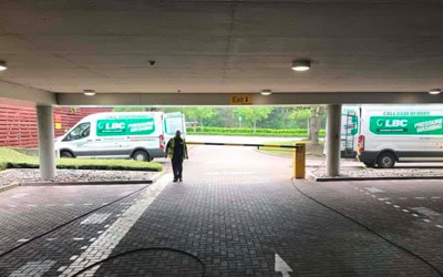 A person wearing high-visibility clothing stands in a parking area beneath a ceiling, facing two white vans with green “LBC” branding and text. The ground is paved with bricks, and there are yellow bollards near the vans. Surrounding the scene are grass and trees.
