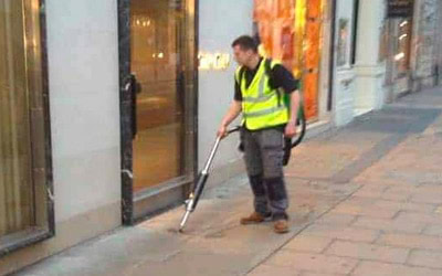 A person wearing a high-visibility vest is cleaning the pavement outside a shop using a backpack vacuum cleaner. The scene takes place on a city street with store windows in the background. The sidewalk is paved with large, rectangular stone tiles.