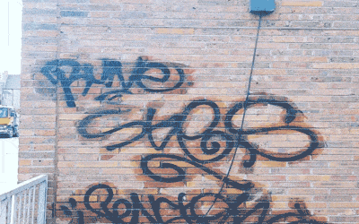 A brick wall with several pieces of black graffiti in various styles and sizes. The graffiti includes large, rounded, and intertwined letters, though they are not easily discernible. There is a blue rectangular object mounted near the top of the wall, with a black cable running down from it.