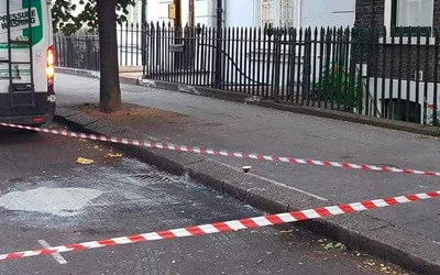 Commercial, A street corner is cordoned off with red and white striped tape. Broken glass is scattered on the ground near the curb. A tree stands on the sidewalk, next to a parked vehicle. A black wrought-iron fence runs alongside a building in the background.