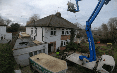 A worker on a blue cherry picker is cleaning the chimney of a two-story house. The house has white walls and a dark tiled roof. The cherry picker extends from a truck with equipment loaded on its bed, parked in the driveway. Trees and neighboring houses are visible in the background.