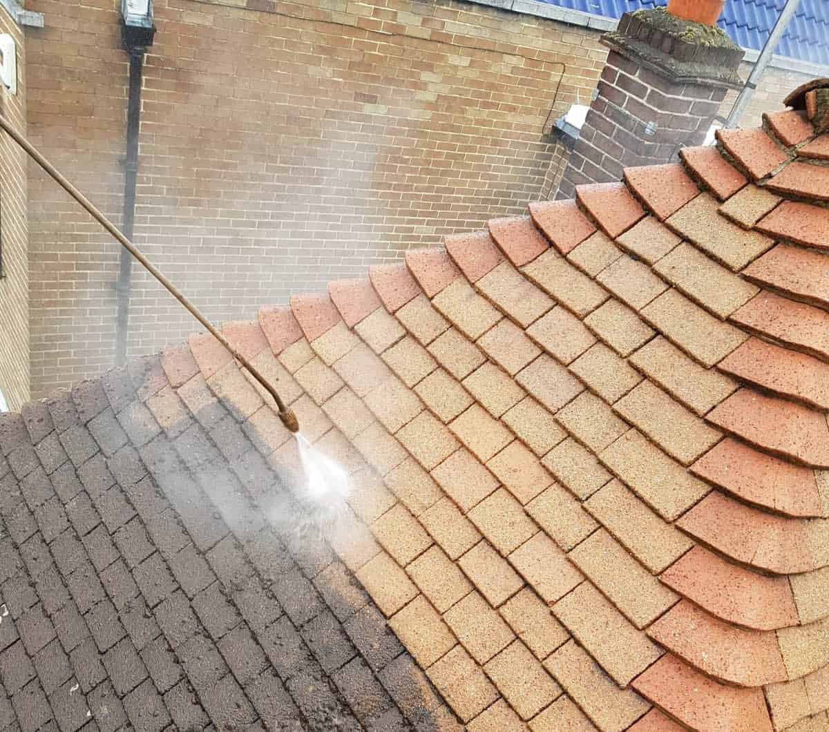 Roof Cleaning review, A roof is being power-washed, with one side visibly darker and dirtier than the other side which appears cleaner. A high-pressure hose sprays water onto the dirty shingles, removing dirt and grime. The roof is composed of interlocking reddish-brown tiles, and brick walls are in the background.