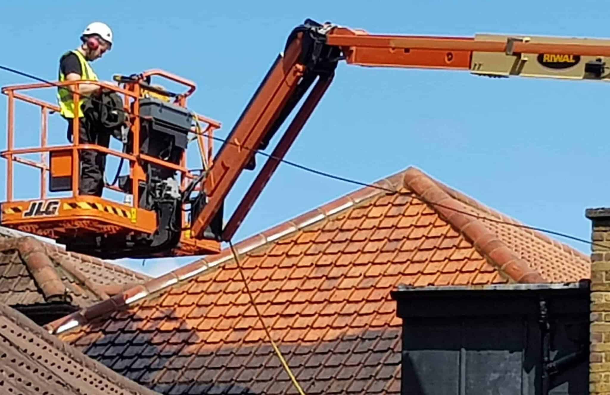A worker, wearing a safety helmet and hi-vis vest, stands on an elevated platform lifted by a JLG boom lift. The platform is positioned above the rooftops of several buildings covered in orange tiles. A blue sky is in the background, with part of the machinery labeled "Riwal" visible.