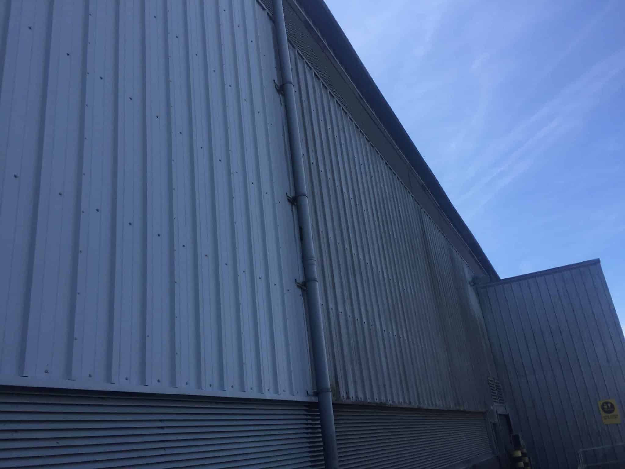 The image shows the side of a large industrial warehouse with corrugated metal siding. There is a drainpipe running vertically along the side of the building. The sky above is blue with wispy clouds. There is a small section of another similar building visible to the right.