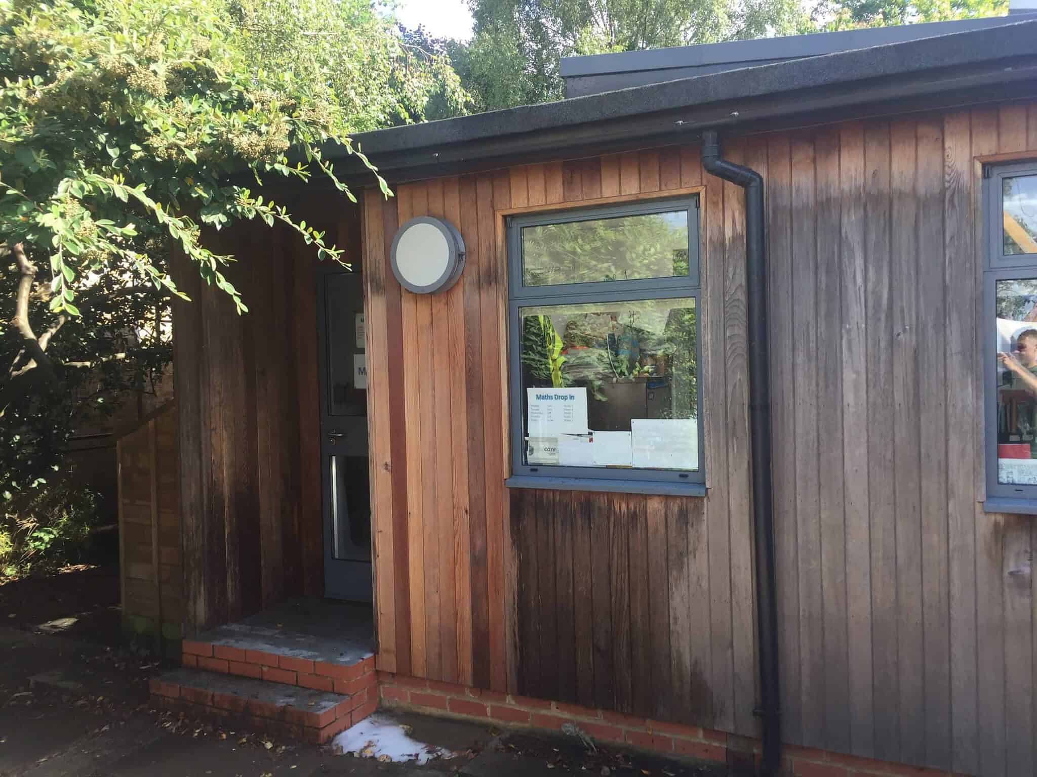 A small wooden building with a modern design featuring vertical wooden planks of varying shades. There is a door to the left with a small set of brick steps leading up to it. Two rectangular windows, partially open, are on the right. Surrounding foliage casts shadows on the building.