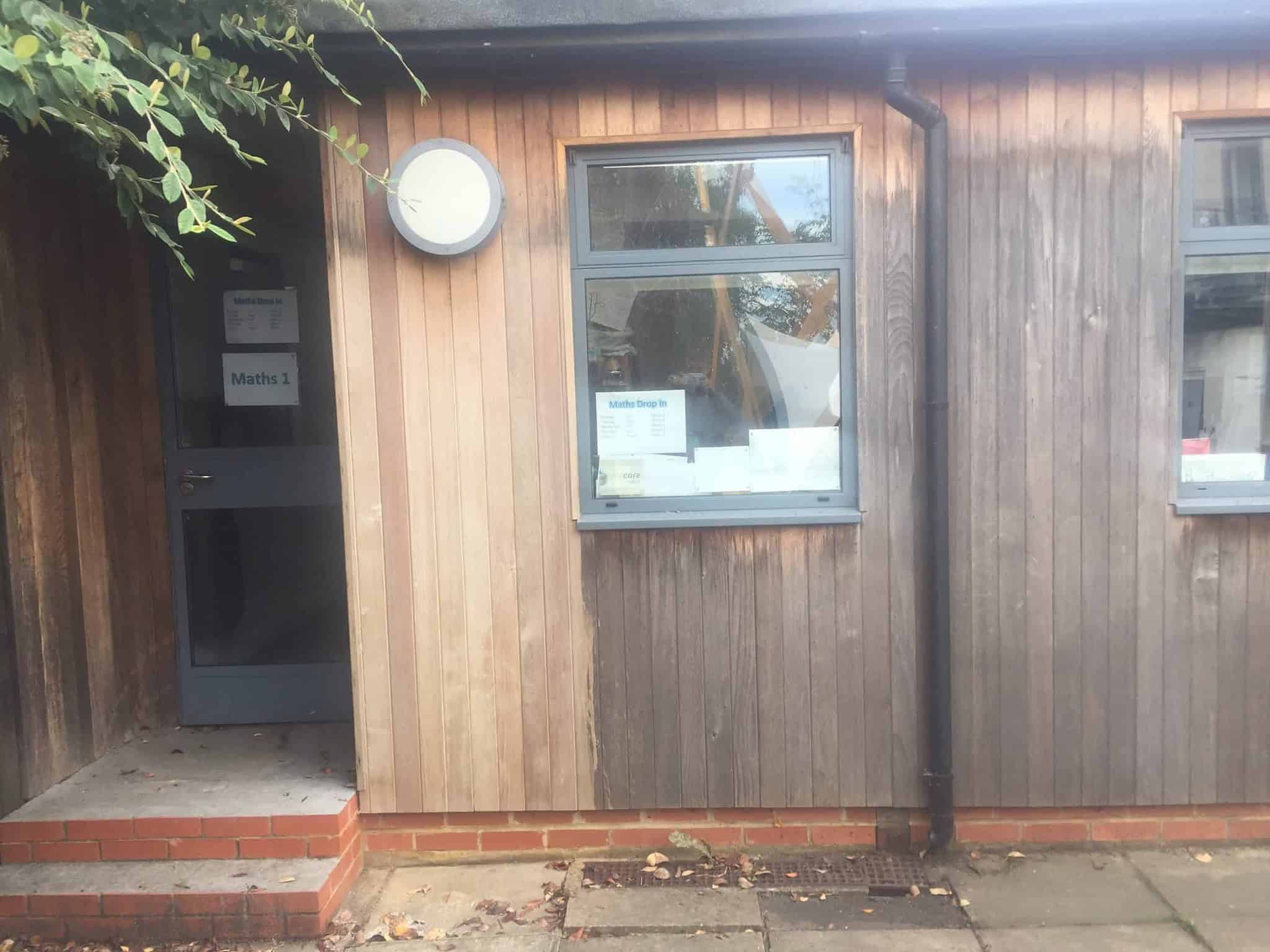 A wooden building featuring a grey door with a "Maths 1" sign and a nearby round wall lamp. Two adjacent windows display "Maths Drop-In" posters. The entrance, with two brick steps, is partially under a leafy branch on the left. The weathered wood shows signs of aging and exposed brick foundation.