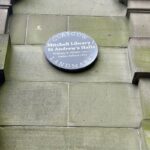 Circular plaque on a stone wall reads: "Glasgow Landmark, Mitchell Library, St. Andrew's Halls, William B. White 1877, James Sellars 1877." The plaque is gray with white text and is set against large, rectangular stone blocks of a building's exterior.