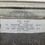 Plaque on a stone wall with text reading: "This stone was laid by Andrew Carnegie, L.L.D., 17th September 1907, The Hon William Bilsland, L.L.D., Lord Provost." The plaque is set in a section of weathered gray stone with small scattered debris on the ground.