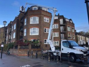 A truck with an extended cherry picker is parked on a street beside a four-story brick building with several windows and balconies. Two workers are on the cherry picker, performing maintenance at the top corner of the building. A bicycle is parked on the sidewalk near a streetlamp.