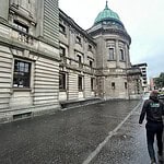 A person in a black hoodie walks on a wet sidewalk beside an old stone building with a green-domed roof. The sky is overcast, and there's a grassy area with trees and parked vehicles on the right. Urban structures are visible in the background.