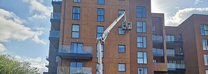 A worker in a cherry picker is inspecting the exterior of a modern apartment building with a mix of wood and brick facade. The building features several glass balconies. The sky is partly cloudy, and the sun is shining from the top right corner, casting shadows.