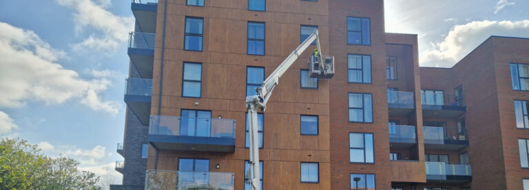 A worker in a cherry picker is inspecting the exterior of a modern apartment building with a mix of wood and brick facade. The building features several glass balconies. The sky is partly cloudy, and the sun is shining from the top right corner, casting shadows.