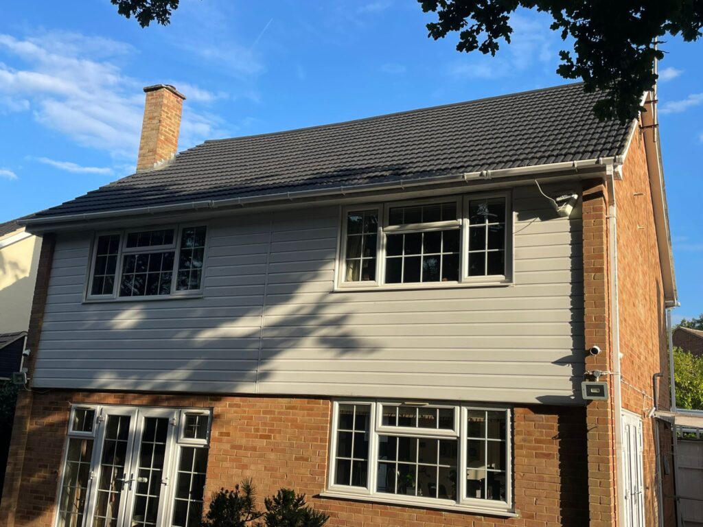 A two-story house with beige horizontal siding on the upper floor and red brick below features white-framed windows, a glass patio door, and a dark gray roof—ideal for Roof Spraying & Recoating Services. Tree branches cast shadows while a chimney stands to the left.