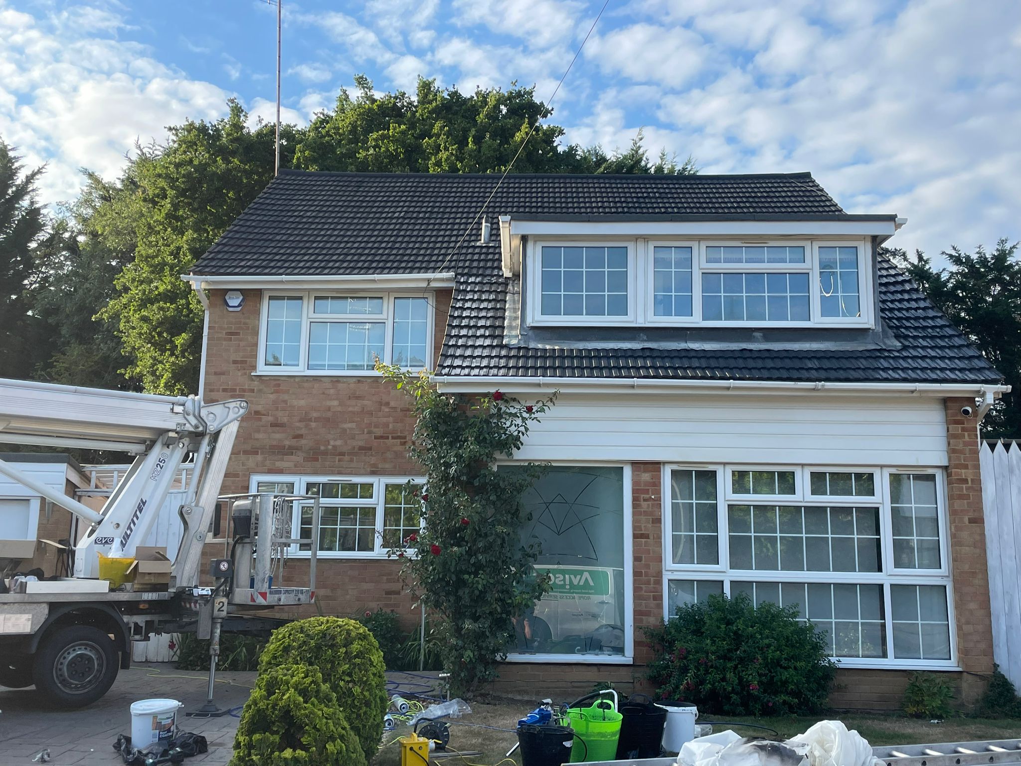 Two-story brick house with large white-framed windows and dark roof tiles. A work truck advertising Roof Spraying & Recoating Services is on the left, with tools and buckets on the lawn. Shrubs and a flowering vine grow in front under a partly cloudy sky.